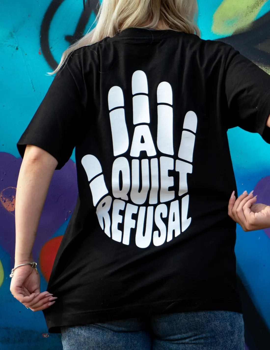 A woman with her back to the camera in front of a graffiti wall, wearing A Quiet Refusal in black.