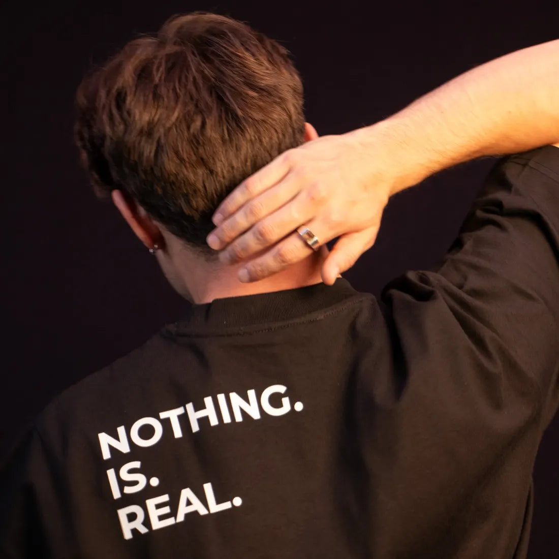 A man with his hand on his neck. The back of his black shirt reads NOTHING. IS. REAL.