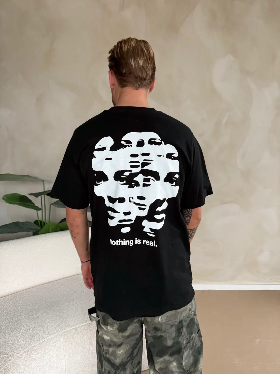 De rug van een zwart shirt met een print van in elkaar overlopende gezichten en nothing is real.